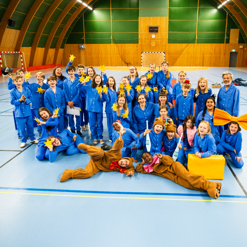 Mikael Holgersson barn och vuxna iklädda blå pyjamasar, hållandes i gula stjärnor står i en sporthall. Två bär björnkostymer
