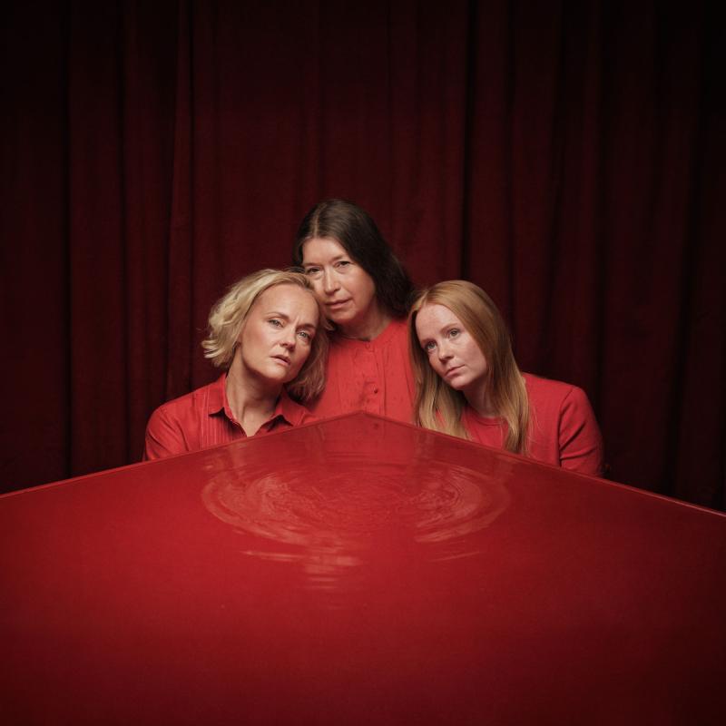 Tre kvinnor vid ett bord. Deras ansikten speglas i den röda bordsskivan.