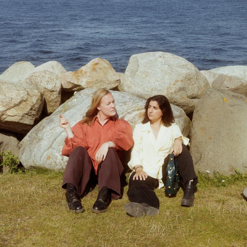 Moa Lundqvist och Dilan Apak sitter på gräset vid stora stenar med havet i bakgrunden