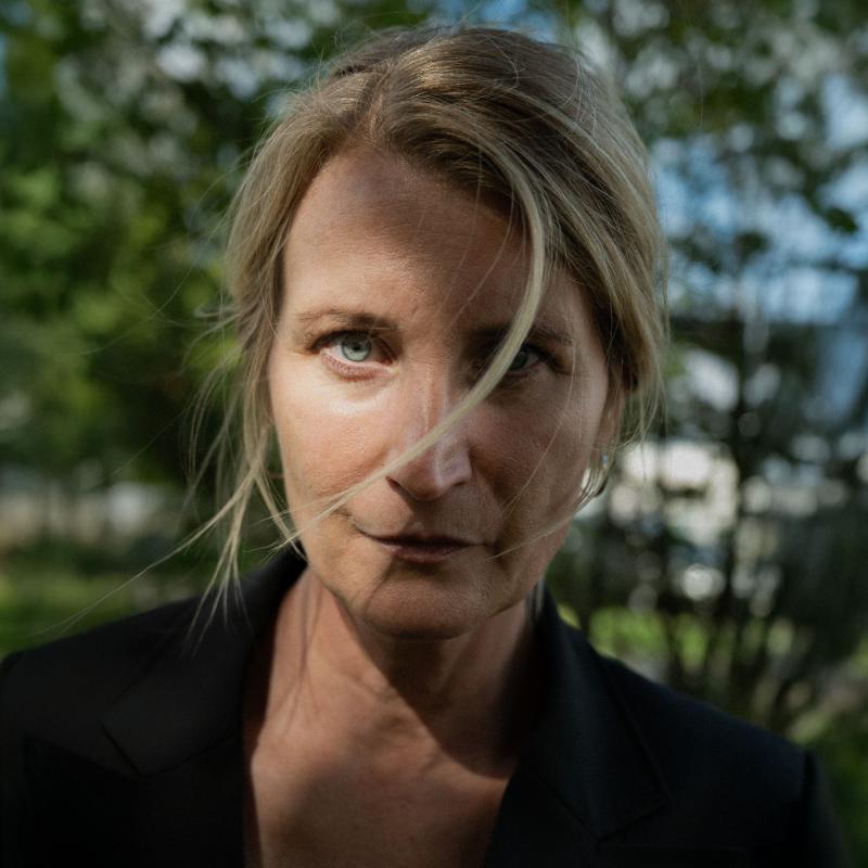 Monica Wilderoth ser in i kameran, en hårslinga framför en ögat, gröna löv i bakgrunden