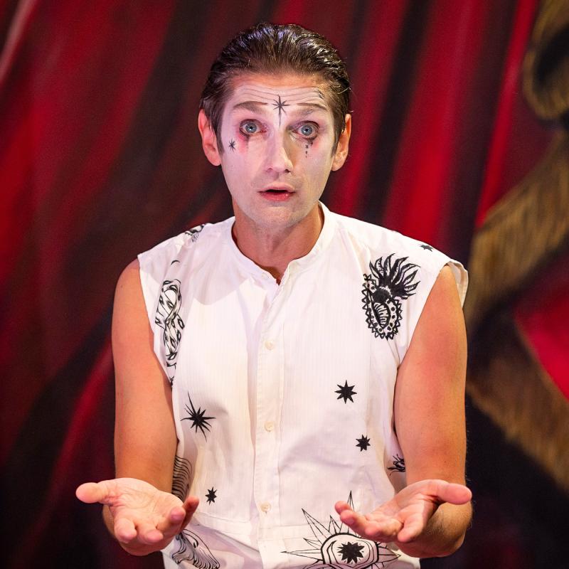 Person med bakslickat hår, vitmålad med svarta mönster i ansiktet ser frågande ut in i kameran