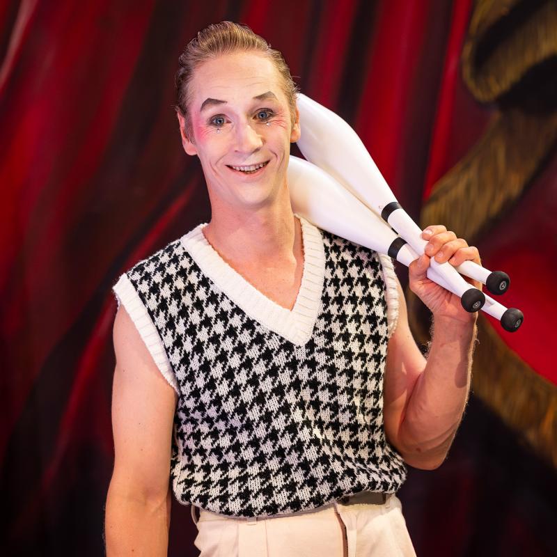 Person med vitsminkat ansikte och rosiga kinder håller i jongleringsklubbor och tittar glatt in i kameran