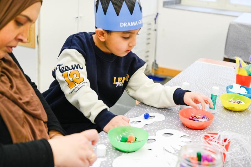 en kvinna och ett barn sitter vid ett bord med teatermasker och paljetter och pysslar