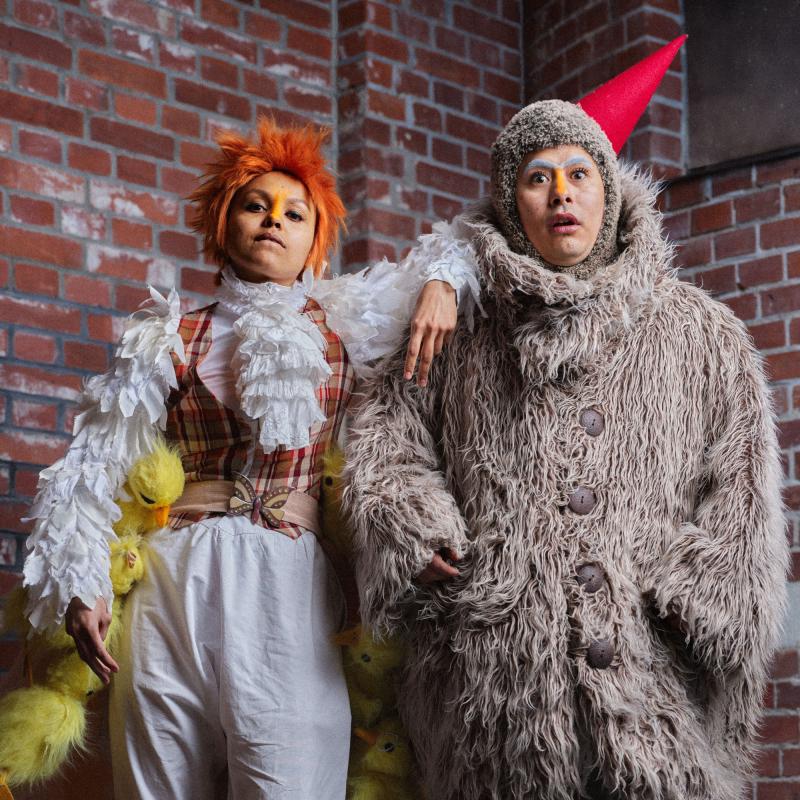 Två personer står mot en tegelvägg - en i grå pälskostym och näbbliknande strut på huvudet, en med vitt krås, vita fjädrar och små gosedjursankungar längs med benen.