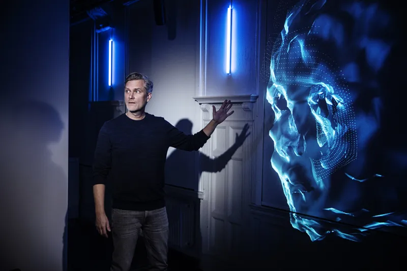 En person tittar på något utanför bild och pekar mot en digital projicering av ett ansikte