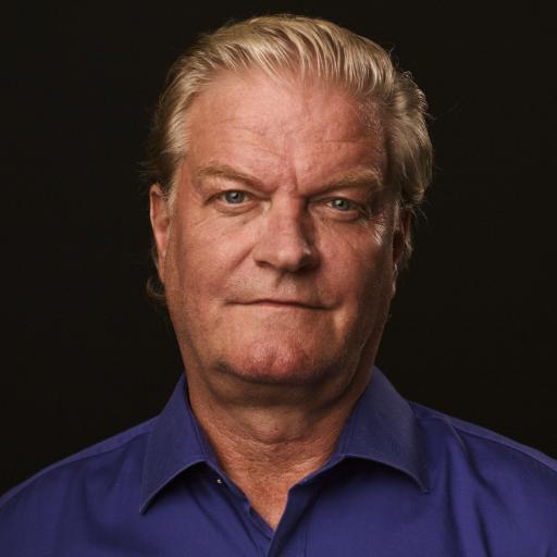 Porträttbild, äldre person med kort blont hår. Skjorta i kobolt blå syns sticka upp. 