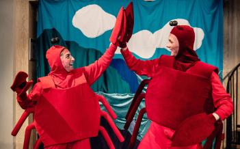Två personer i krabbkostymer: röda overaller med krabbskal, röda mössor med antenner och krabbklor på händerna. Gör high five