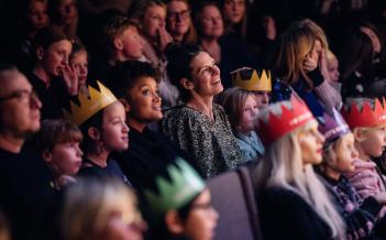 publik i salong som tittar rakt fram, barn och vuxna blandat. Några skrattar andra tittar fokuserat. En del har papperskronor på huvudet.