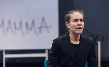 Skådespelare till höger i kavaj sitter ner med kraftfullt ansiktsuttryck. Ordet Mamma står på väggen bakom.