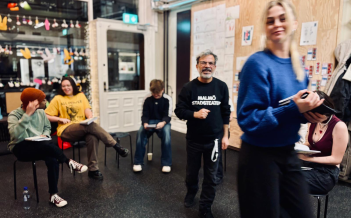 En workshopmiljö med människor i samtal och aktivitet. En äldre man i en Malmö Stadsteater-tröja står i mitten och ler mot kameran, medan andra deltagare sitter på stolar i bakgrunden, pratandes eller skrivandes. En kvinna i blå tröja rör sig framåt med en anteckningsbok i handen.