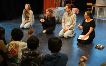 En grupp barn sitter på golvet och lyssnar uppmärksamt på fyra vuxna som också sitter på golvet i en cirkel på en teaterscen. De vuxna ser engagerade ut; en av dem håller i en anteckningsbok och en penna. Scenografin i bakgrunden inkluderar en konstgjord sten, en röd vägg och rekvisita som ger en rustik känsla. Ett av barnen räcker upp handen.