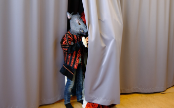 Anders Andersson Ett barn med svartrödrandig kostymkappa och en mask i form av ett vildsvinshuvud står halvt gömd bakom en ridå