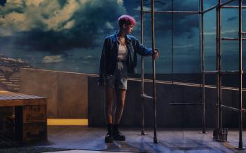 En person med jeansjacka, jeansshorts, rosa kort hår och svarta boots står på en scen med stålstativ och en himmelsbakgrund