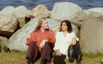 Två personer sitter på gräset lutade mot stenar med havet bakom sig.