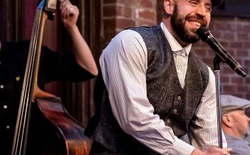 Jenny Baumgartner Person med skjorta, väst och keps står vid mikrofon och kontrabas i bakgrunden